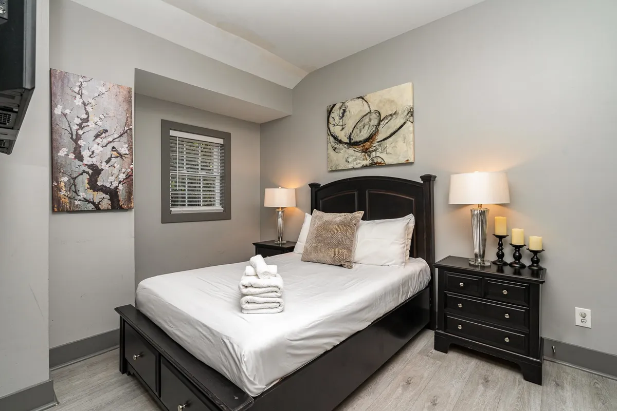 A queen bed dressed in soft white linens, accented with decorative pillows and modern artwork on the walls. A nightstand with lamps adds to the inviting, restful feel, highlighting a cozy, intimate environment.