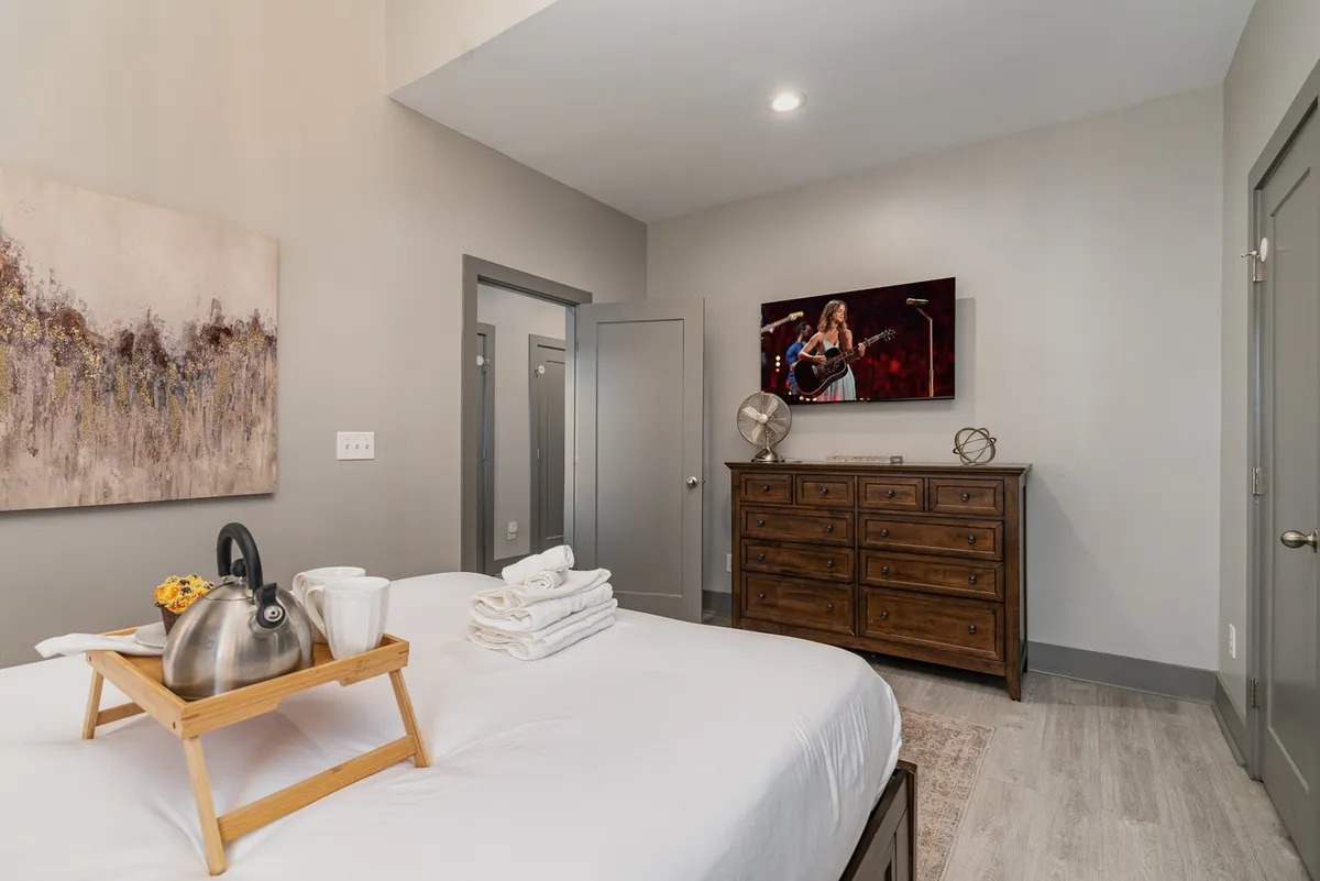 A cozy queen bedroom featuring a bed with crisp white linens, a wooden dresser, a wall-mounted television displaying a performance, tasteful artwork above the bed, and a breakfast tray with a kettle and cups, all illuminated with soft, inviting light.