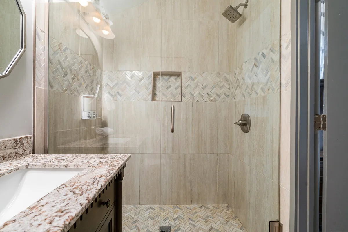A stylish bathroom with a granite countertop, modern shower fixtures, and a patterned tile design. The space is bright and clean, suggesting a spa-like experience and the presence of quality bath essentials.
