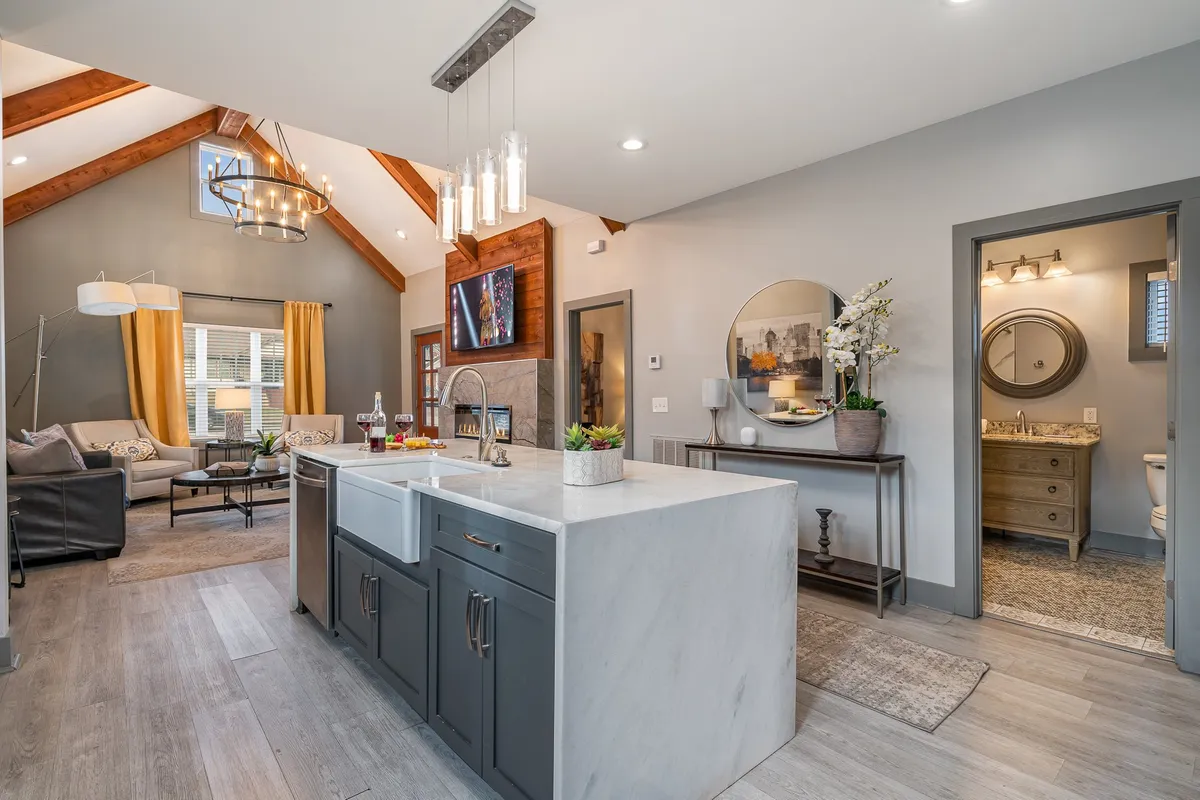A spacious open-concept living and kitchen area featuring a stylish kitchen island with a sink, plush living room seating, large windows streaming in natural light, and a statement chandelier. The seating area and kitchen are visually connected, creating a welcoming space for gatherings and relaxation.