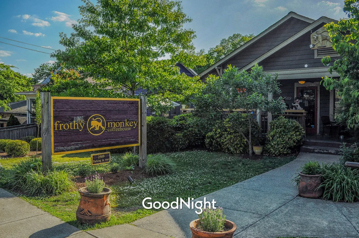 This image captures the exterior of the Frothy Monkey coffeehouse, with its inviting sign, lush green foliage, and a welcoming pathway leading to the entrance. The scene exudes a cozy, neighborhood feel and suggests a vibrant, local gathering spot.