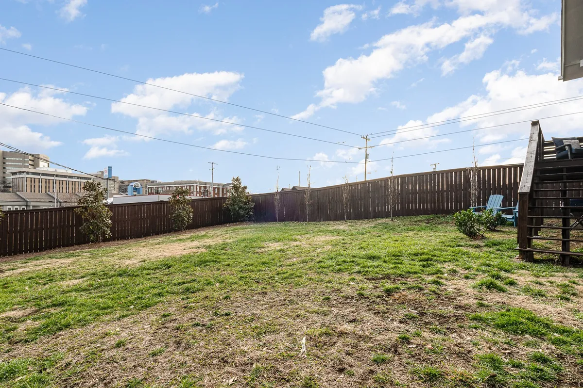 A wide view of an expansive backyard lawn bordered by a wooden fence and adjoining buildings. The space is open, green, and offers ample room for children to play, families to gather, or pets to explore. The mood is relaxed and the area is bathed in natural light, suggesting a safe and welcoming environment for outdoor activities.