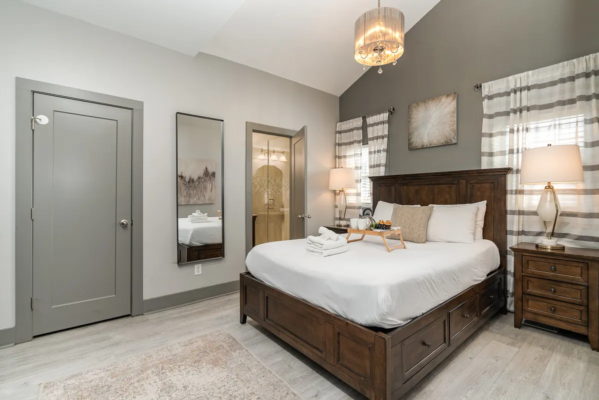 A serene queen bedroom highlighted by a bed with white linens, a wooden nightstand topped by a glowing lamp, decorative wall art, and a mirror reflecting warm natural light. The overall mood is peaceful and perfect for unwinding.