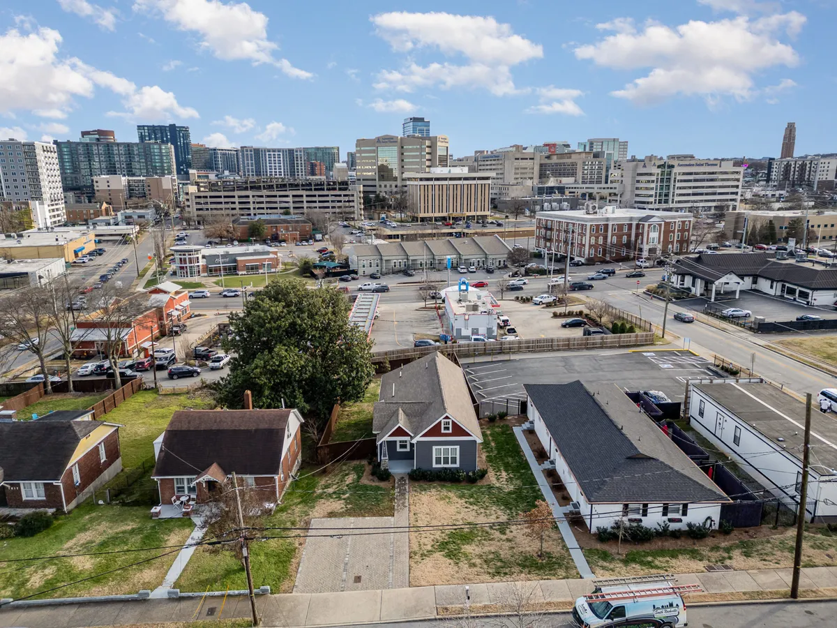 A view over a welcoming neighborhood with residential houses, green lawns, and a glimpse of the city skyline in the background. The setting suggests a vibrant, community-oriented environment with space for gatherings and local events, blending residential charm with city access.
