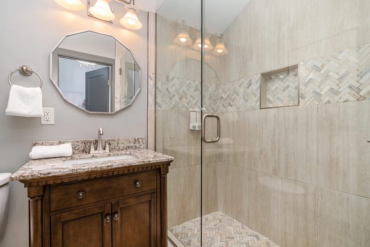 A stylish bathroom featuring a granite countertop, a glass shower enclosure with a herringbone tile design, modern light fixtures, and a decorative mirror. The space feels refreshing and spa-like, ideal for morning or evening routines.