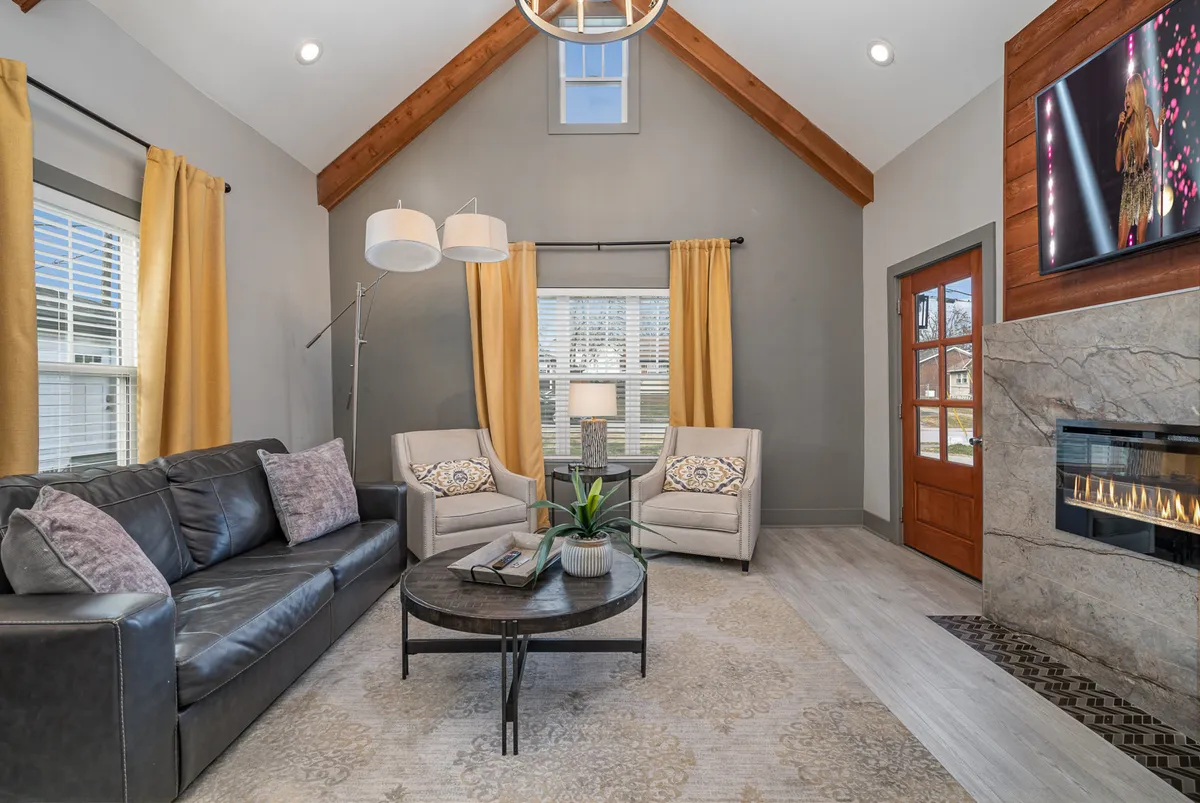 A cozy living room scene with a black leather sofa, a warm fireplace, a large coffee table, expansive windows, and a dramatic chandelier overhead. The arrangement invites guests to gather, relax, and enjoy shared moments by the fire or while watching TV.