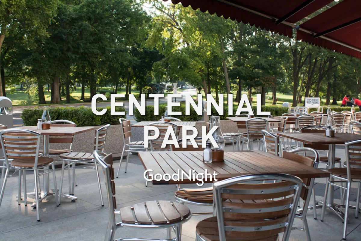 A cheerful outdoor patio scene with tables and chairs under a canopy, surrounded by lush park greenery. The setting is ideal for socializing, dining al fresco, or simply unwinding in a lively community space.