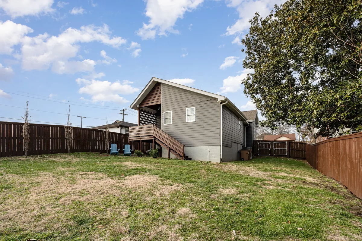 A sunlit backyard featuring an open lawn with lawn chairs, stairs leading up to a porch, and a relaxed, cheerful atmosphere. The space is clearly designed for group enjoyment, casual gatherings, and outdoor activities.